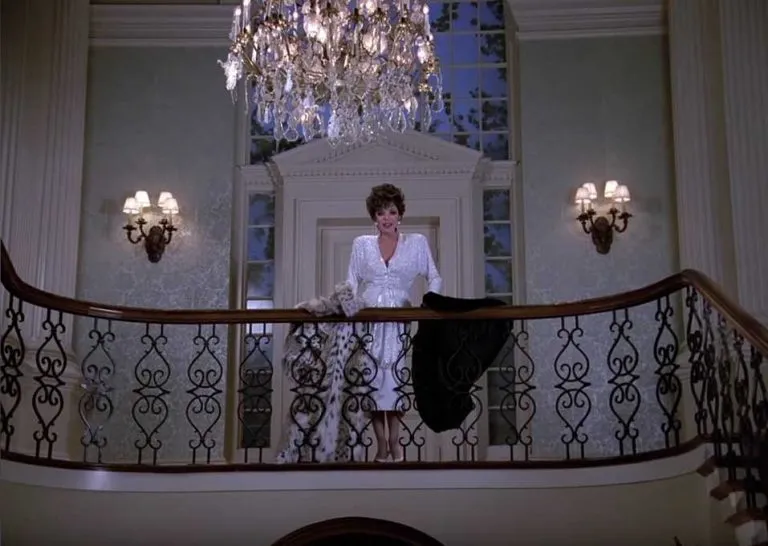 Alexis Carrington overlooking the foyer from the grand stairway where she's draped a fur coat over the handrail.